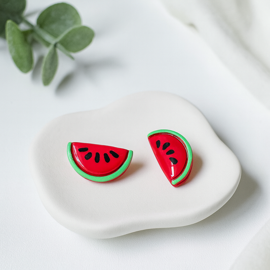 Watermelon slice earrings on a white stone surface with a leafy branch in the background